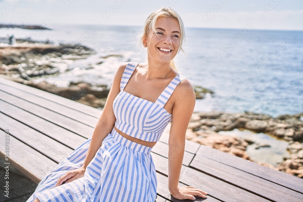 Poster Young blonde girl smiling happy sitting on the bench at the beach.