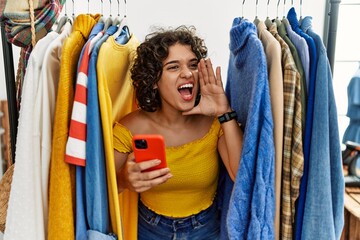 Obraz na płótnie Canvas Young hispanic woman searching clothes on clothing rack using smartphone shouting and screaming loud to side with hand on mouth. communication concept.