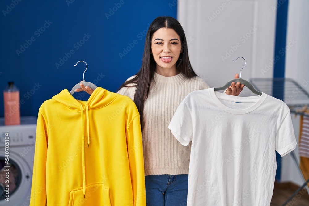 Poster young woman hanging clothes on racks sticking tongue out happy with funny expression.