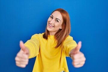 Fototapeta premium Young woman standing over blue background approving doing positive gesture with hand, thumbs up smiling and happy for success. winner gesture.