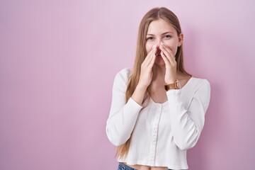 Young caucasian woman standing over pink background laughing and embarrassed giggle covering mouth...