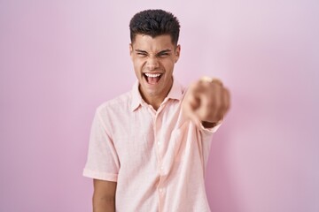 Young hispanic man standing over pink background pointing displeased and frustrated to the camera,...