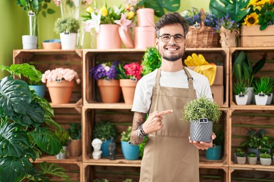 Young hispanic man with tattoos working at florist shop holding plant smiling happy pointing with hand and finger
