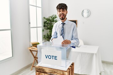 Hispanic man with beard voting putting envelop in ballot box sticking tongue out happy with funny...