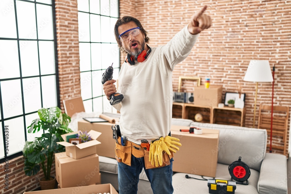 Wall mural Handsome middle age man holding screwdriver at new home pointing with finger surprised ahead, open mouth amazed expression, something on the front