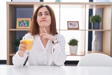 Brunette woman drinking glass of orange juice thinking concentrated about doubt with finger on chin and looking up wondering