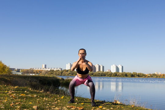 A young woman in a tracksuit squats during a workout in a park against the background of the city.