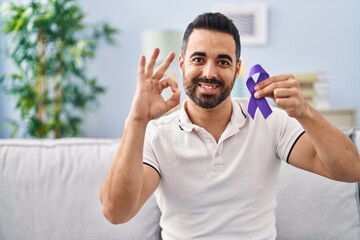 Young hispanic man with beard holding purple ribbon awareness doing ok sign with fingers, smiling...