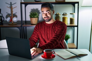 Young hispanic man with beard using computer laptop at night at home smiling looking to the side and staring away thinking.