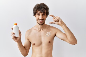 Fototapeta premium Young hispanic man standing shirtless holding sunscreen lotion smiling and confident gesturing with hand doing small size sign with fingers looking and the camera. measure concept.