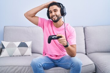 Hispanic young man playing video game holding controller sitting on the sofa smiling confident...