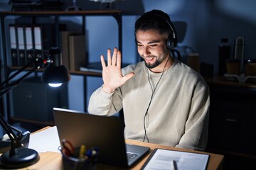 Young handsome man working using computer laptop at night showing and pointing up with fingers number five while smiling confident and happy.