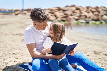 Father and daughter reading book sitting on sand at beach