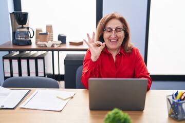 Senior woman with glasses working at the office with laptop doing ok sign with fingers, smiling friendly gesturing excellent symbol