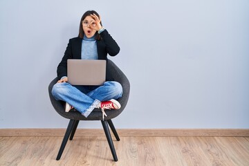 Young hispanic woman sitting on chair using computer laptop doing ok gesture shocked with surprised...