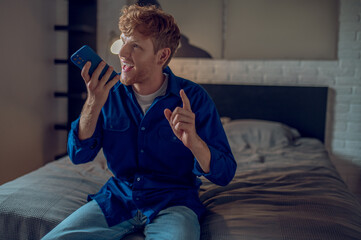 A young man in dark-blue shirt sitting on the bed and having a call - obrazy, fototapety, plakaty