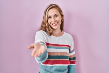 Obraz premium Young blonde woman standing over pink background smiling cheerful offering palm hand giving assistance and acceptance.