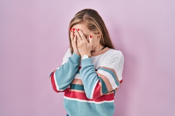 Young blonde woman standing over pink background with sad expression covering face with hands while...