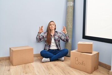 Young blonde woman sitting on the floor moving to a new home relax and smiling with eyes closed doing meditation gesture with fingers. yoga concept.