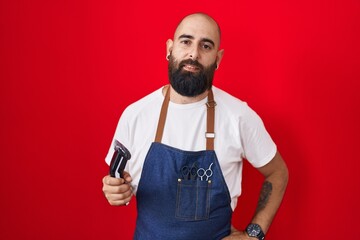 Young hispanic man with beard and tattoos wearing barber apron holding razor relaxed with serious expression on face. simple and natural looking at the camera.