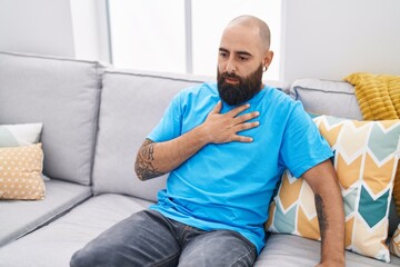 Young bald man sitting on sofa suffering heart attack at home