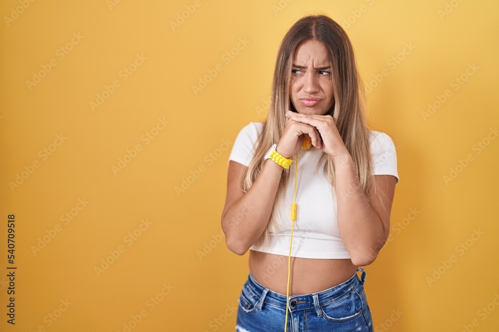 Poster young blonde woman standing over yellow background wearing headphones laughing nervous and excited w