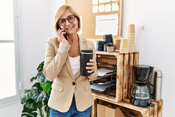 Middle age blonde woman talking on the smartphone drinking coffee at office