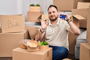 Plus size hispanic man with beard moving to a new home holding credit card doing ok sign with fingers, smiling friendly gesturing excellent symbol