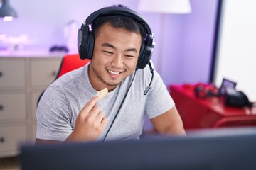 Young chinese man streamer playing video game eating chips potatoes at gaming room