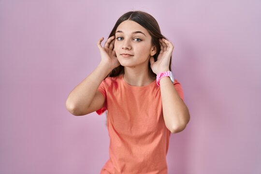 Teenager girl standing over pink background trying to hear both hands on ear gesture, curious for gossip. hearing problem, deaf