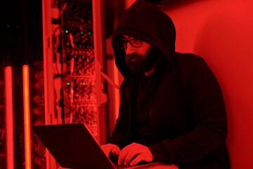 Hacker wearing hoodie doing coding on laptop in server room