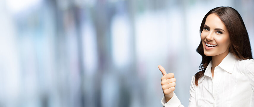 Portrait of very happy smiling young excited businesswoman, showing thumb up hand sign gesture. Success in business concept. Cheerful woman standing on blurred office background.
