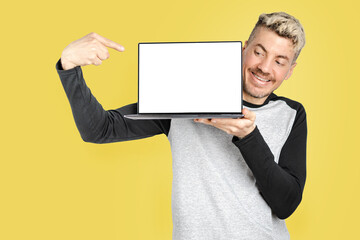 Young adult man wearing casual clothing holds open laptop and points with finger at white screen, mock up for copy space and advertising. Isolated studio yellow background