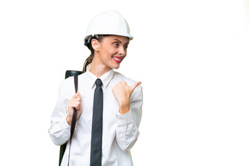 Young architect woman with helmet and holding blueprints over isolated background pointing to the side to present a product