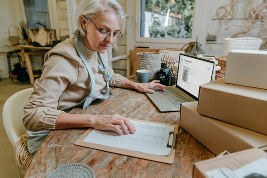Craftswoman reading checklist sitting with laptop on workbench