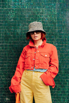 Hipster fashion young woman in bright clothes, sun glasses and bucket hat posing on the green tile wall background. Urban city street fashion. Vertical card. Selective focus. Copy space.