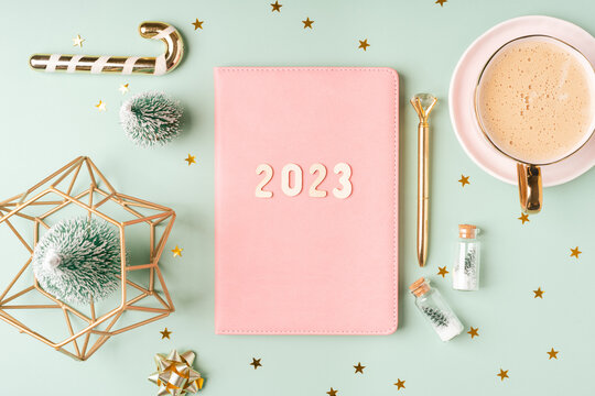 Christmas pink notepad with 2023 letters text. Flat lay on green mint table background with planner, cup of coffee, candle, Christmas decoration, notebook and stationery. Top view Desktop 