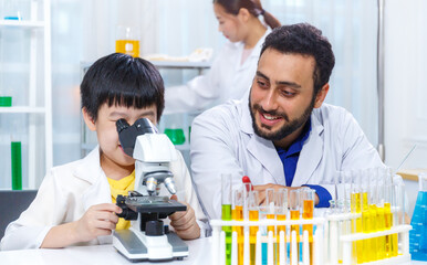 A male teacher sits at a classroom desk with a male student using a microscope while learning a...