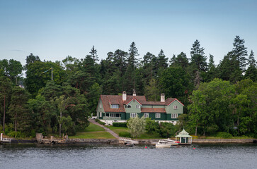 Fototapeta premium Sweden, Stockholm - July 16, 2022: Morning approach by ship from Baltic. Green mansion on Ostra Granholmen island north shore west of Edlunda ferry terminal. Green foliage and blue sky