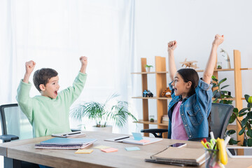 Fototapeta premium excited kids showing win gesture near notebooks and paper notes on table at home.
