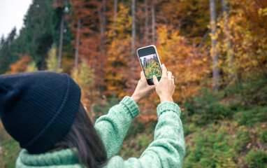 A young woman in a black hat takes a photo of the autumn forest on a smartphone.