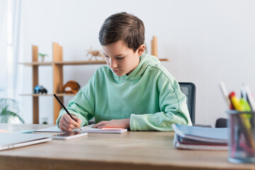 Naklejka na ściany i meble focused boy doing homework and writing in copybook near blurred mobile phone.