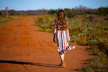 a beautiful long-haired girl in a long dress walks along a road in the desert in western australia,...