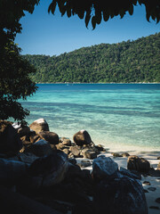 Scenic view of Sunset Beach, Koh Lipe Island. White sand beach, clear turquoise sea with coral reef transparent water against summer blue sky, tree and rocky coastline foreground. Satun, Thailand.