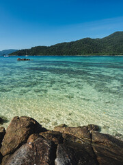 Scenic view of Sunset Beach, Koh Lipe Island. Crystal clear shallow water with coral reef transparent against summer blue sky, rocky coastline and Koh Adang Island background. Satun, Thailand.