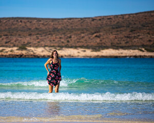 Fototapeta premium beautiful long-haired girl in a dress soaks her legs in turquoise water on a paradise beach at turquoise bay in cape range national park near exmouth, western australia; beach with red cliffs in the b