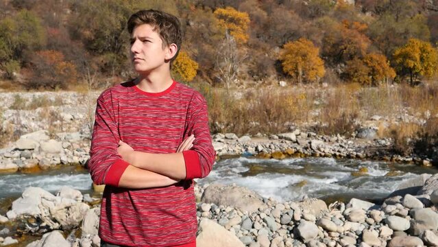 the boy stands in the mountains in front of the river with crossed arms 