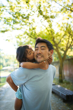 Close-up of Asian father and his daughter spending time in park. Happy young dad holding little kid on his back and girl hugging him kissing his cheek. Resting together and happy childhood concept