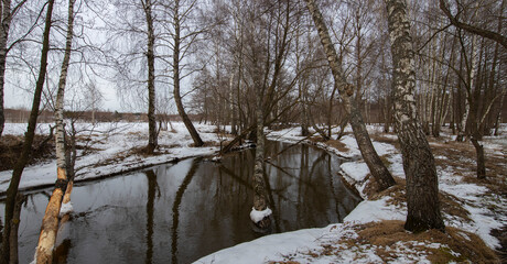 Fototapeta na wymiar Flood. March evening by the river, the snow is melting. Evening in early spring. Trees are reflected in the water.