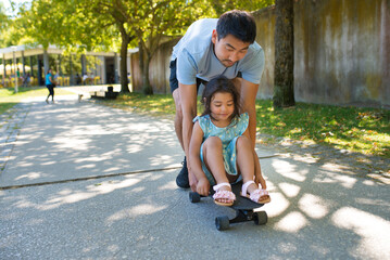 Portrait of Asian dad and girl resting with skateboard in park. Bearded man in casual clothes...